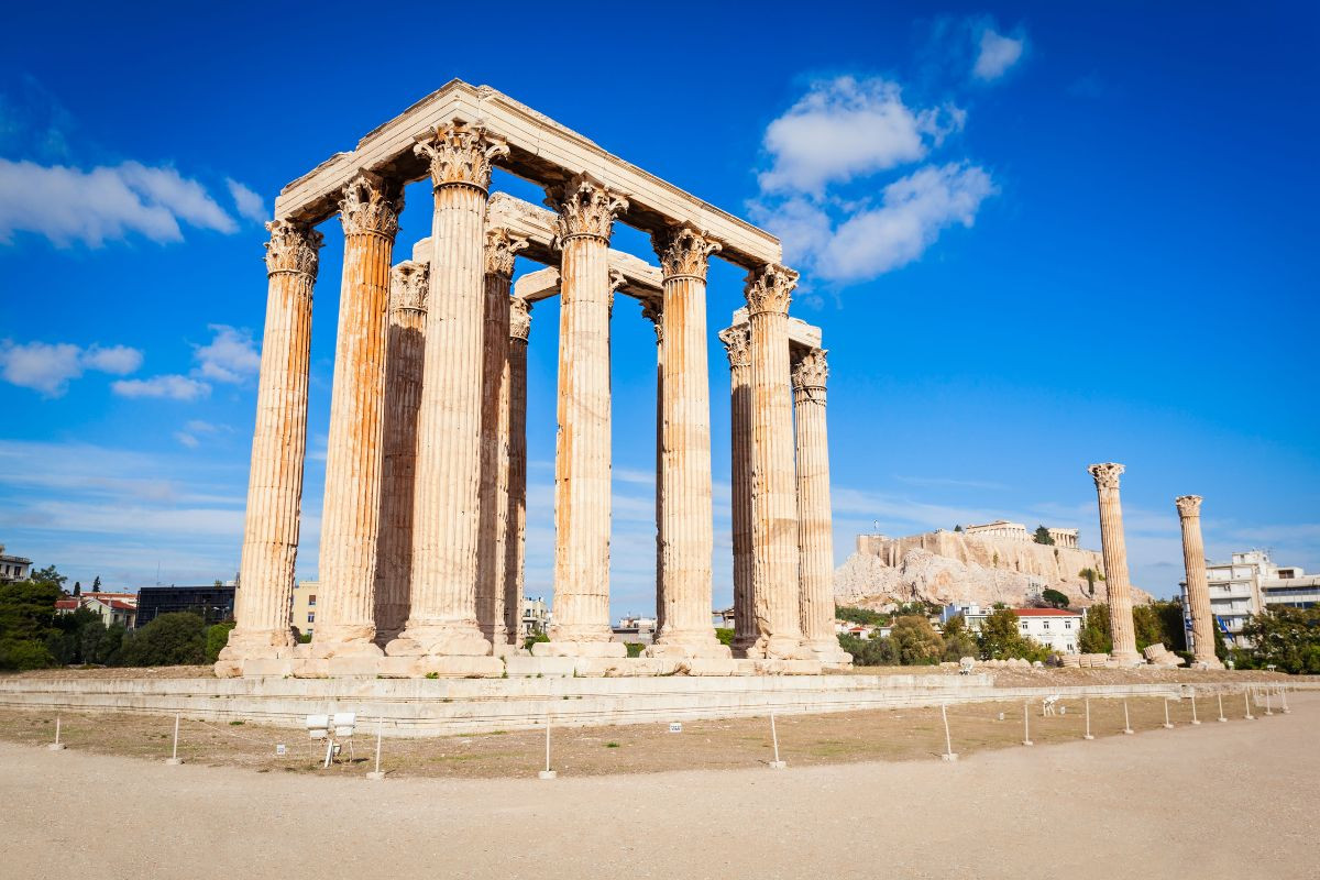 The image shows the Temple of Olympian Zeus in Athens, Greece. Several tall ancient columns remain standing, with a few fallen ones nearby. The Acropolis is visible in the background under a clear blue sky_top 10 places to visit in Athens